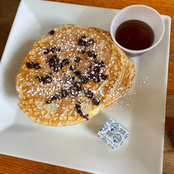 Chocolate chip pancakes with powdered sugar, a side of syrup, and a butter packet on a white plate.