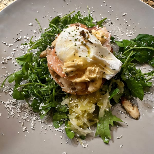 A Gold Standard Cafe breakfast dish with poached eggs, hollandaise, prosciutto, and greens on a gray plate.