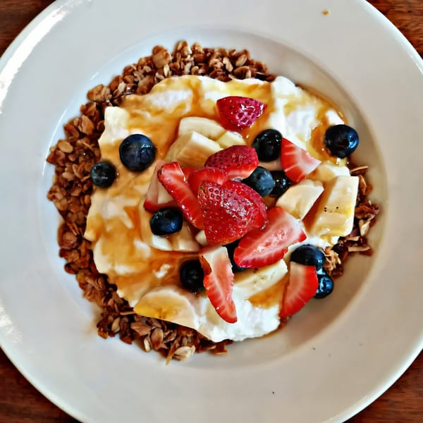 A white bowl of Gold Standard Cafe's granola, yogurt, strawberries, blueberries, and bananas, drizzled with honey.